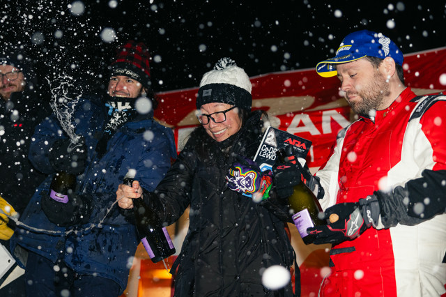 A podium celebration. Multiple contestants are spraying champagne as snowflakes fall.