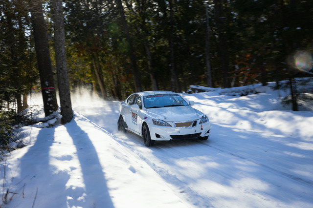 A white Lexus IS250 races around a corner on icy roads in a forest. The car has racing media applied, and the radiator is blocked off with a piece of cardboard.