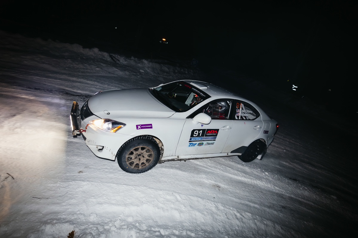 A white Lexus IS250 slides through an icy turn at night. It has a light bar on the front of the car, and various logos adorning the car sides.