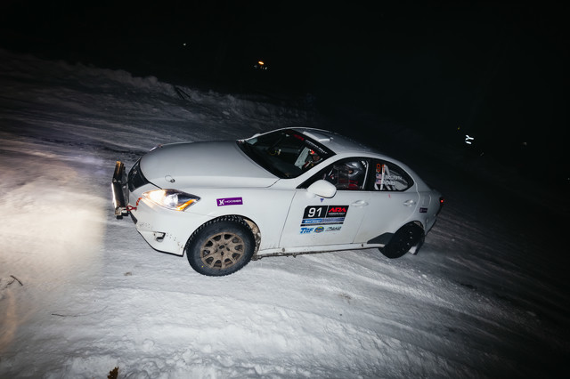 A white Lexus IS250 slides through an icy turn at night. It has a light bar on the front of the car, and various logos adorning the car sides.
