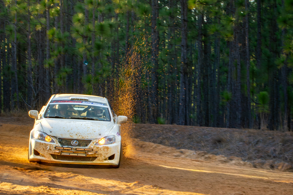 A white Lexus IS250 slides through sand. The nose of the car is weighted down as it brakes, while sand is kicked up in the rear from the wheels spinning.