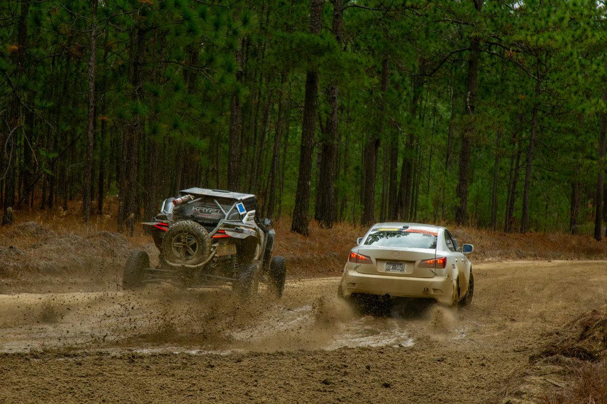 A white Lexus IS250 slides through a corner, being chased by a blue off road vehicle. They are racing through the woods, and the surface of the corner is slippery.