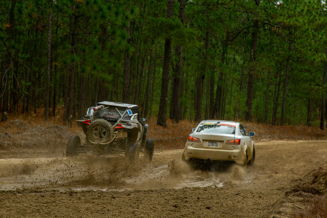 A white Lexus IS250 slides through a corner, being chased by a blue off road vehicle. They are racing through the woods, and the surface of the corner is slippery.