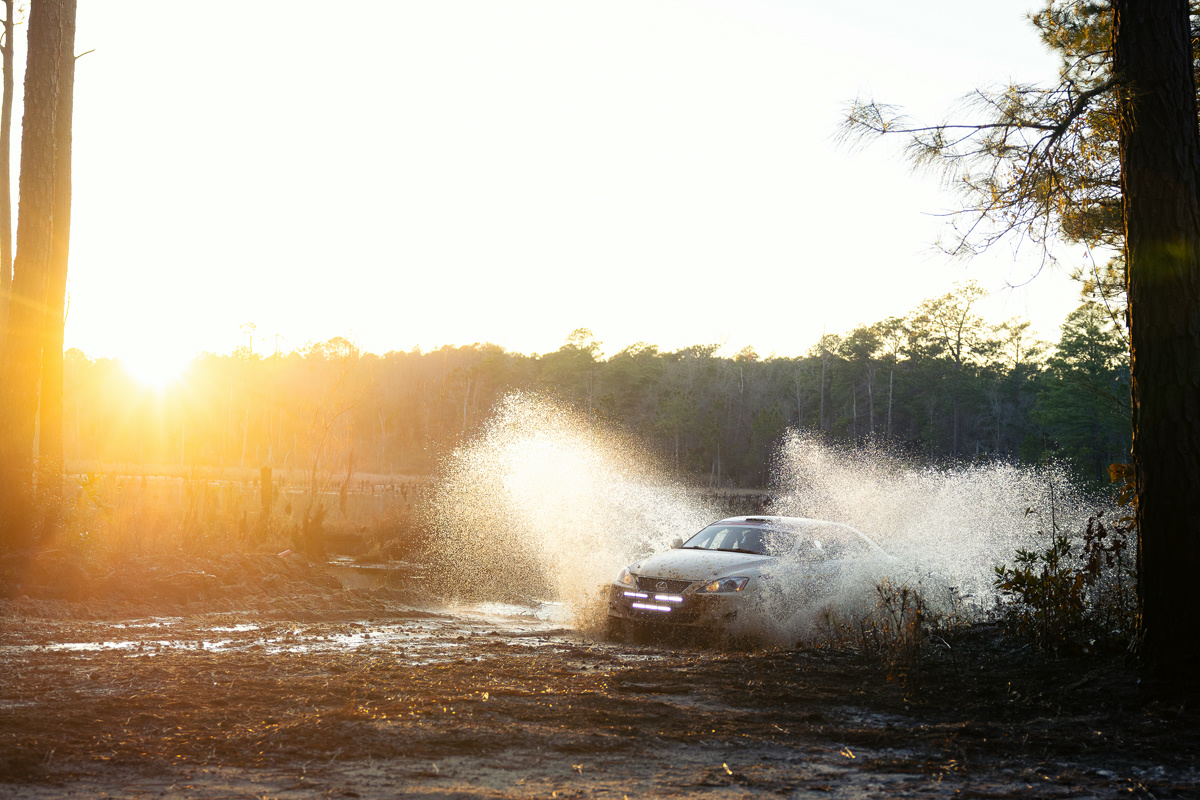 A white Lexus IS250 pushing through a water crossing. A desolate lake can be seen behind.