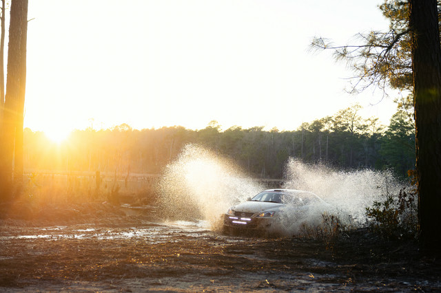 A white Lexus IS250 pushing through a water crossing. A desolate lake can be seen behind.