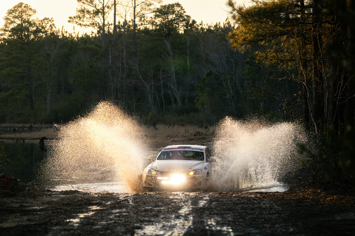 A white Lexus IS250 pushing through a water crossing. A desolate lake can be seen behind.
