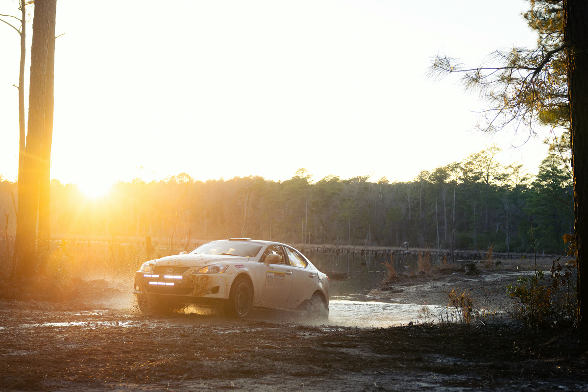 A white Lexus IS250 exiting a water crossing. A desolate lake can be seen behind.