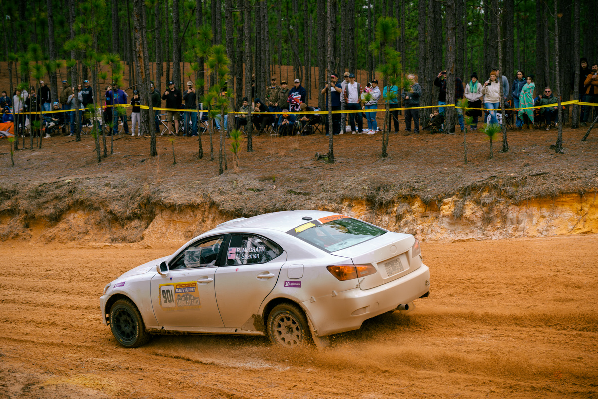 A white Lexus IS250 powers through a sandy turn in the woods. Spectators line the tree bank above.
