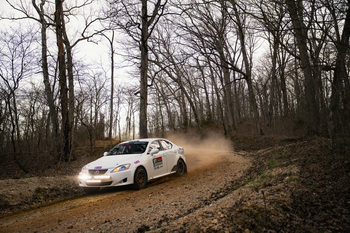 A white Lexus IS250 coming around a corner in thee woods.