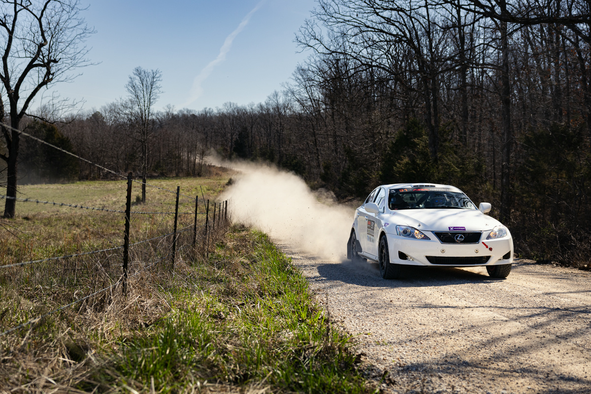 A white Lexus IS250 speeding down a rural road. A long dust cloud is visible in the background.