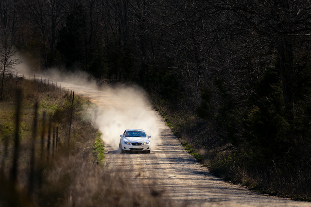 A white Lexus IS250 speeds down a rural road. A dust cloud can be seen in its wake.