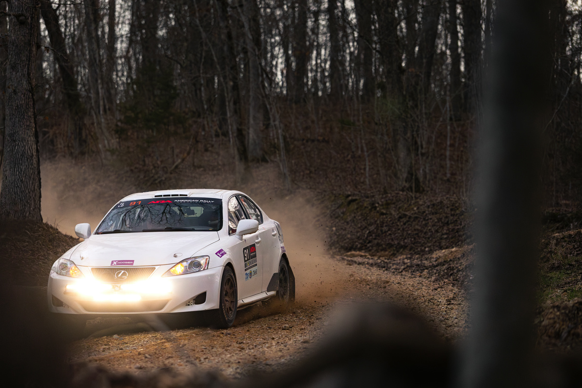 A white Lexus IS250 coming around a corner deep in the woods. The front of the car has an illuminated light bar on it.