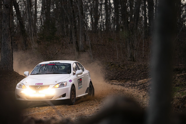 A white Lexus IS250 coming around a corner deep in the woods. The front of the car has an illuminated light bar on it.