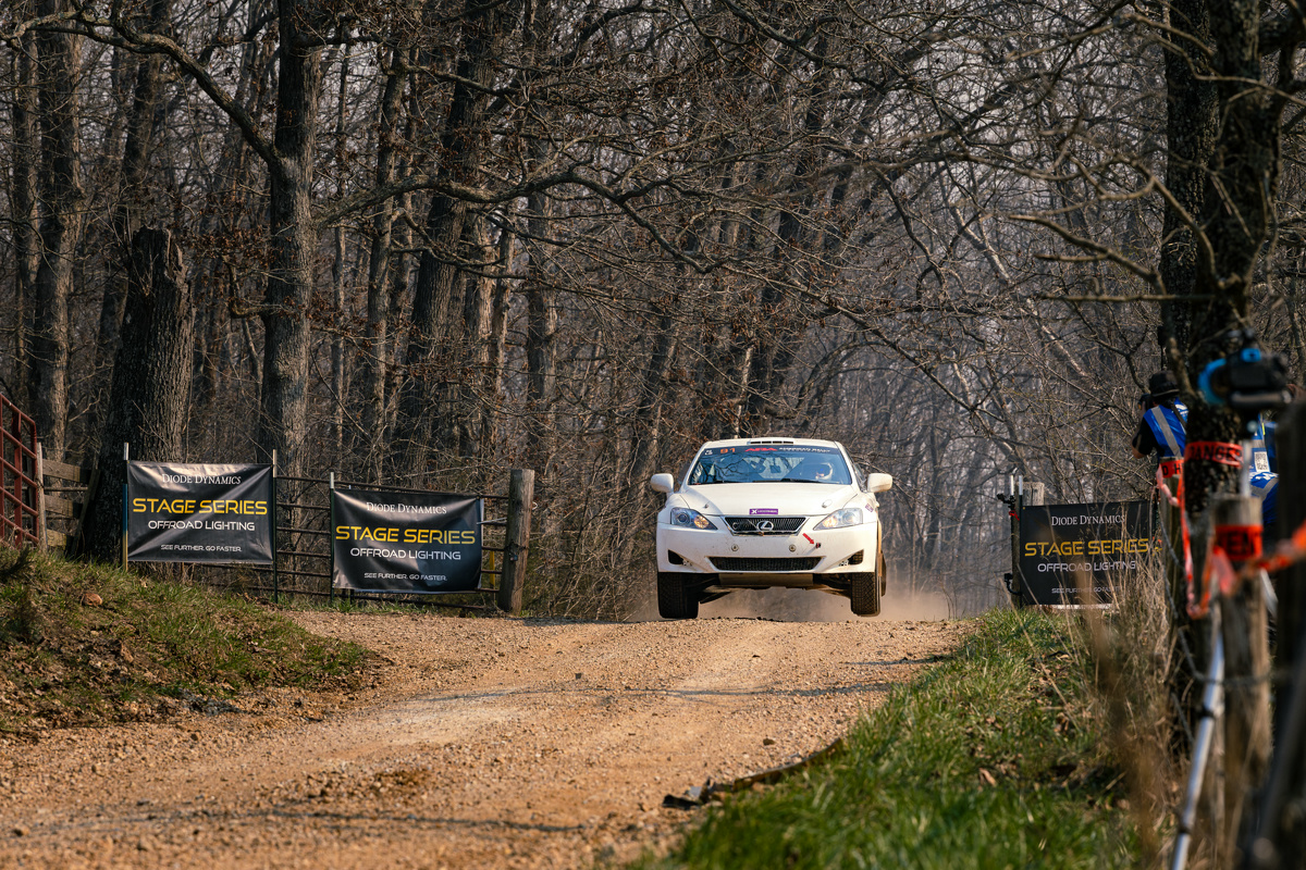 A white Lexus IS250 doing a mini jump over an infamous cattle guard crossing.