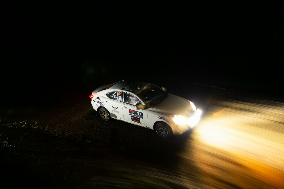 A white Lexus IS250 racing through the woods at night. The lights on the front of the car are intensely bright.