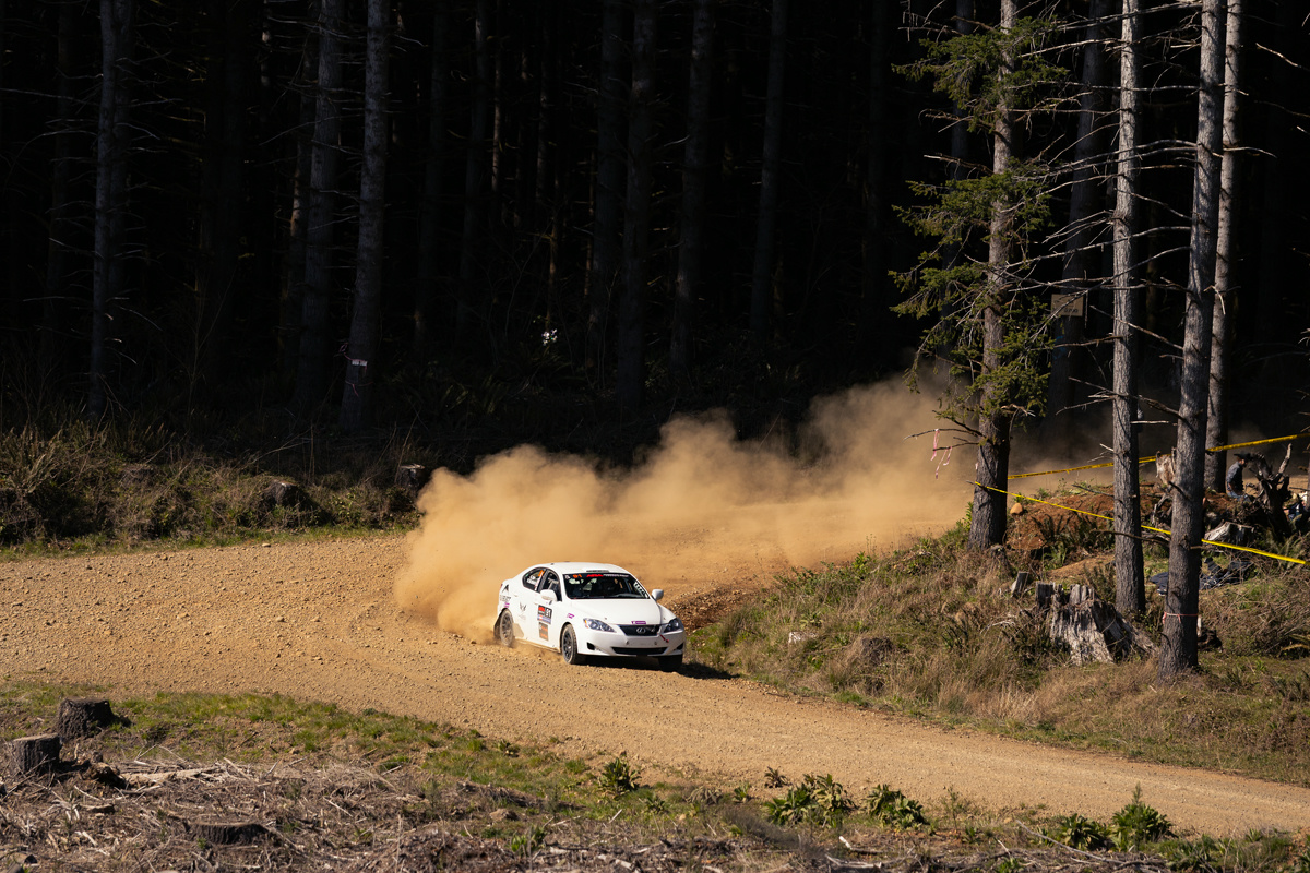 A white Lexus IS250 drifting around a turn. A massive dust cloud can be seen behind it. Yellow banner tape is cordoning off a safe racing space.