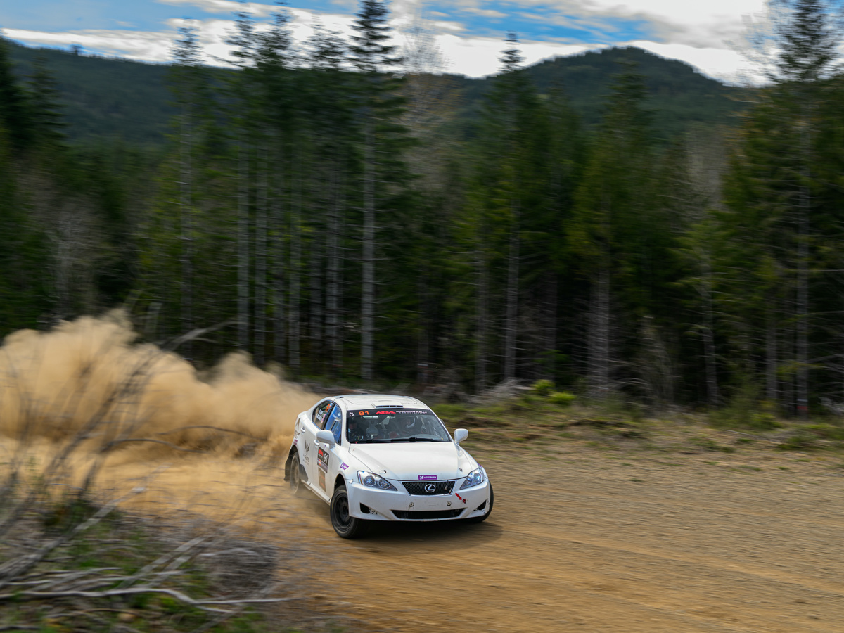 A white Lexus IS250 slides through a turn on a closed logging road. Massive dust can be seen in the wake of the car.