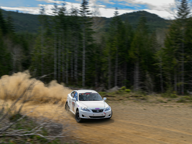 A white Lexus IS250 slides through a turn on a closed logging road. Massive dust can be seen in the wake of the car.