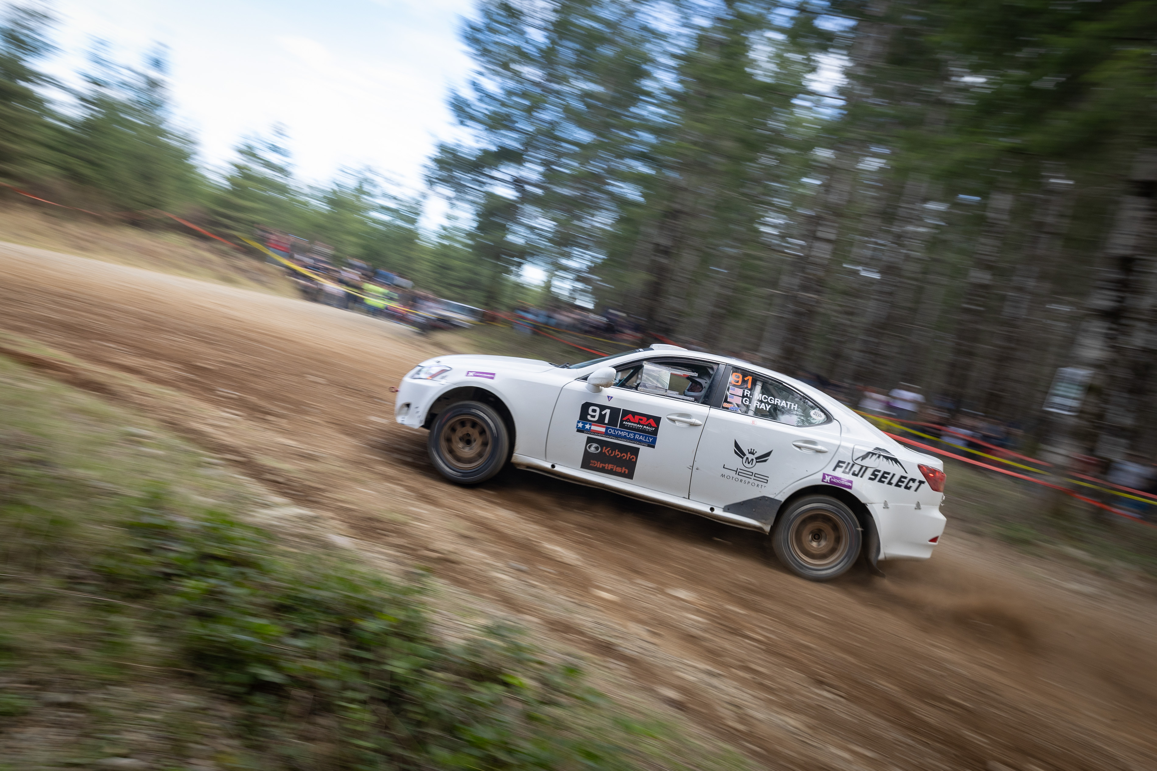 A white Lexus IS250 navigates a hairpin turn in front of a crowd of spectators. Logos adorn the car and dirt is kicked up behind it. The spectators are lined by trees.