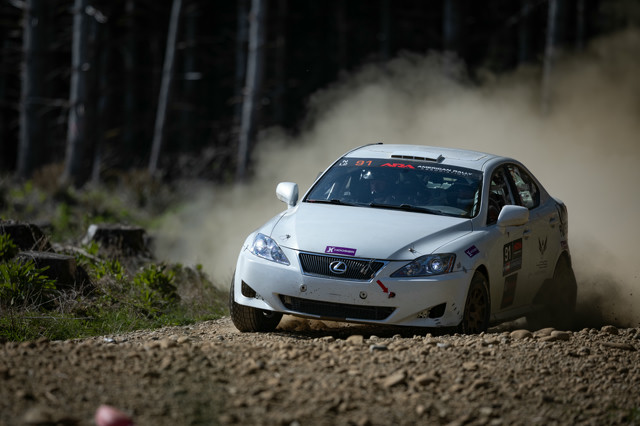 A white Lexus IS250 braking hard before sliding through a dirt turn. The front end is heavily compressed and a massive dust cloud can be seen behind it.