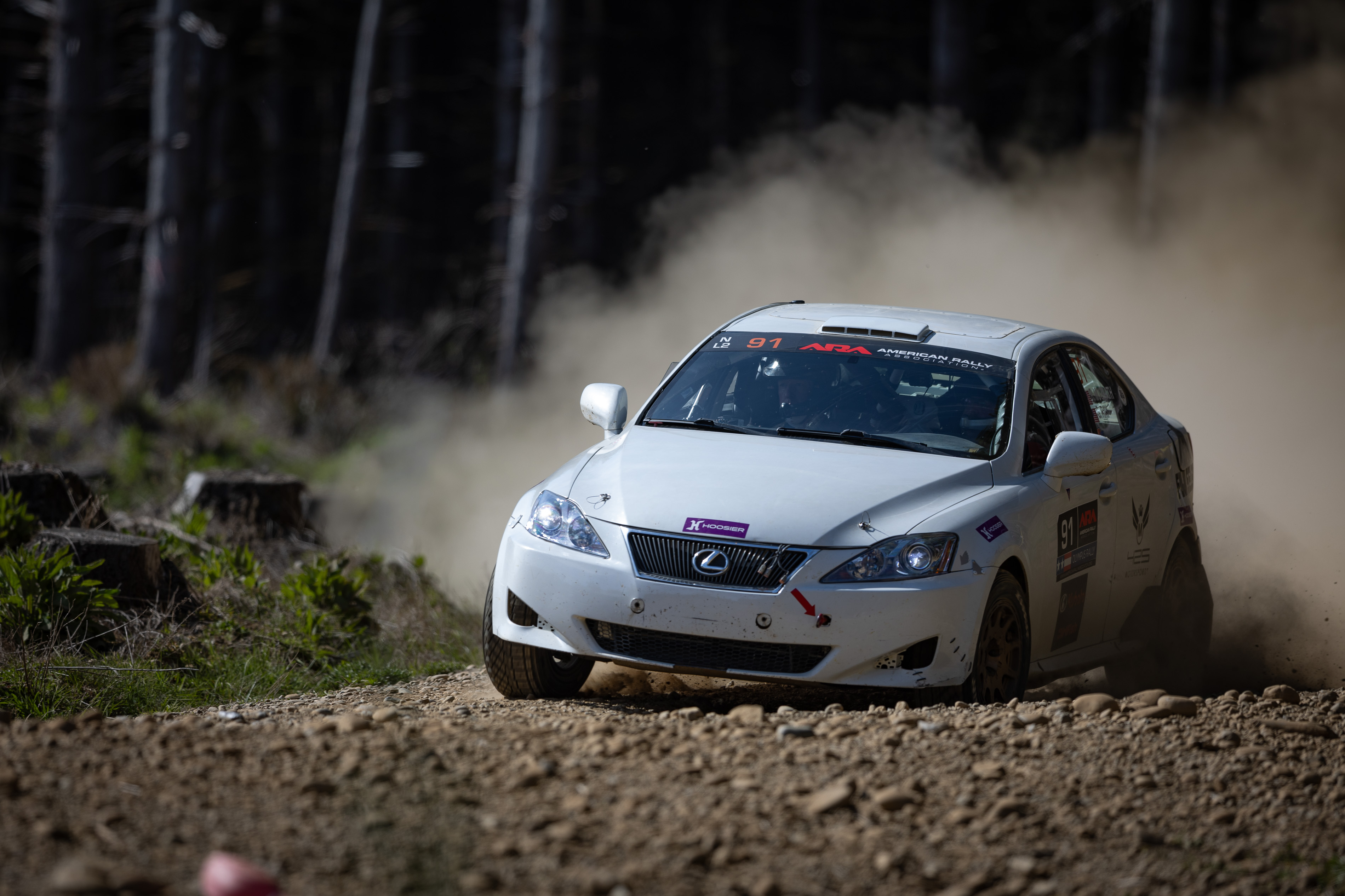 A white Lexus IS250 braking hard before sliding through a dirt turn. The front end is heavily compressed and a massive dust cloud can be seen behind it.
