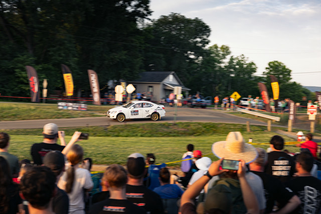 A crowd taking photos of a white Lexus IS250 speeding over a bump in the road.