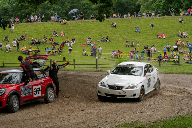 A white Lexus IS250 taking the long way around a Mini Cooper on a dirt road. People in the distance are cheering.