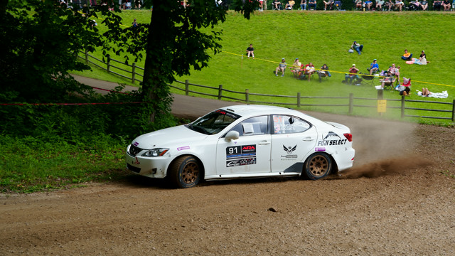 A white Lexus IS250 slides through a turn on gravel. People in the background are watching and cheering. The back end of the Lexus is squatted down, implying heavy throttle usage.