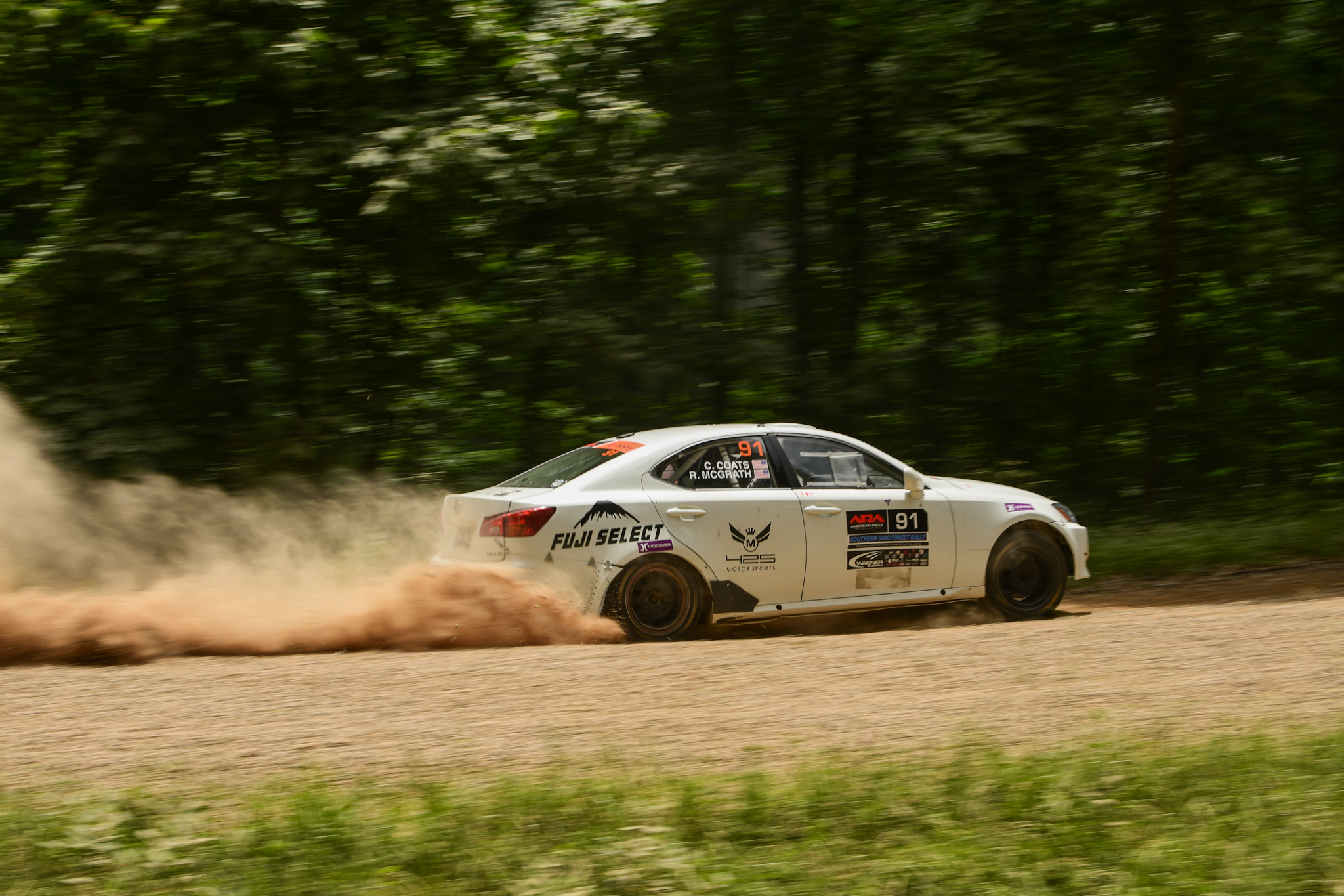 A white Lexus IS250 powering through a turn. It's heavily squatting in the back and massive dust clouds trail it.