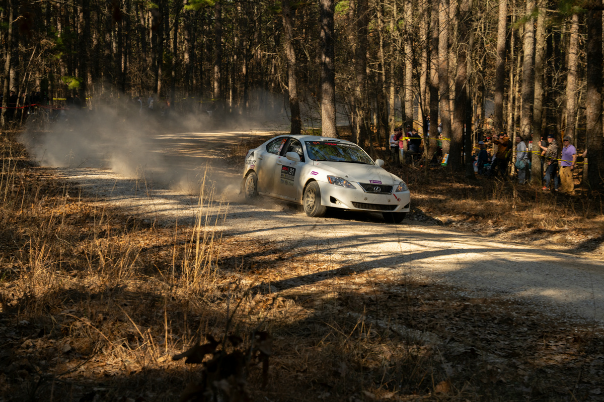A white Lexus IS250 blasts through a dirt road as onlookers gaze from the trees.