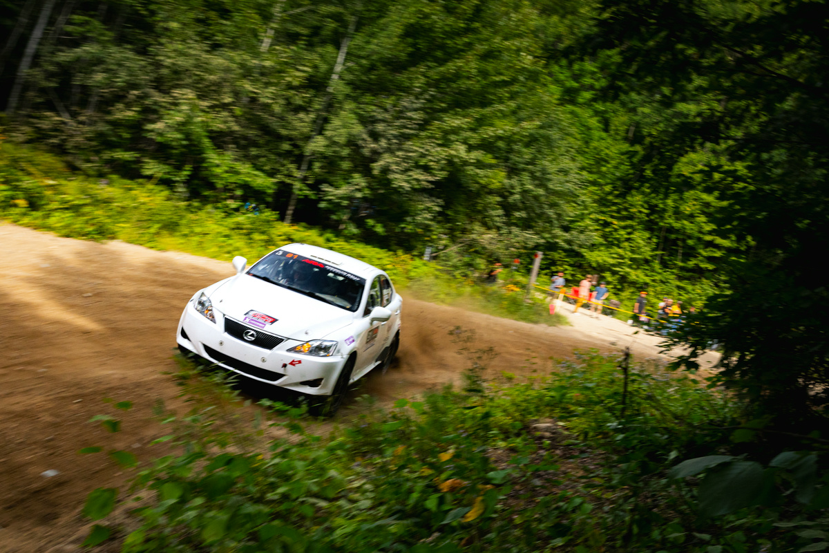 A white Lexus IS250 screaming through a turn in the forest. An onlooking crowd can be seen behind.