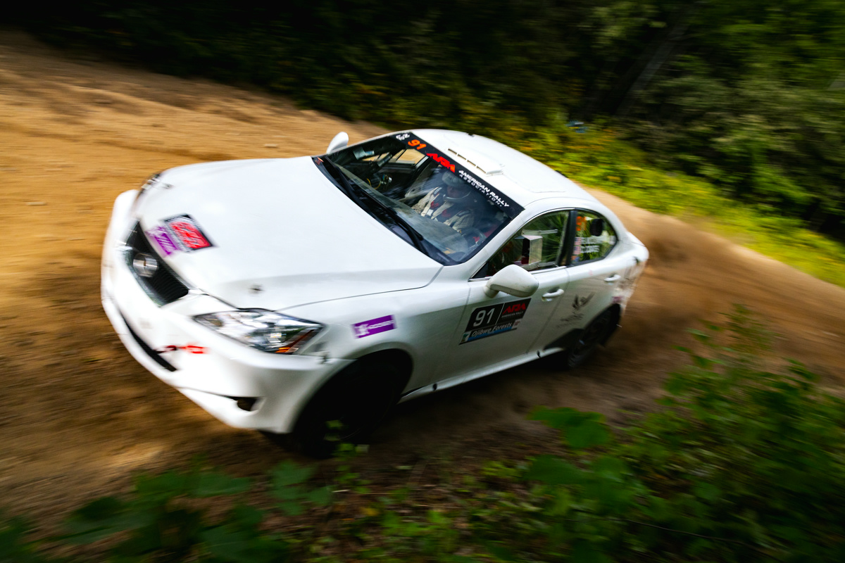 A white Lexus IS250 mid-corner in the forest. The co-driver is visible in the car and calling notes.