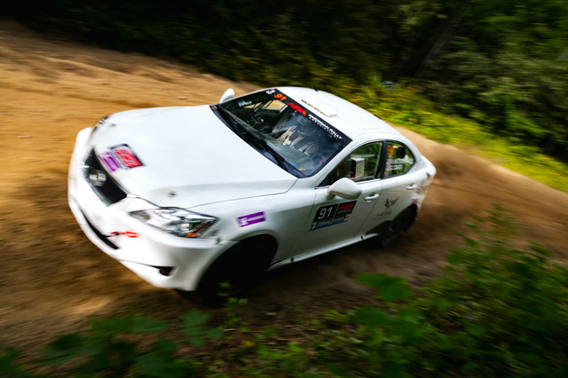 A white Lexus IS250 mid-corner in the forest. The co-driver is visible in the car and calling notes.