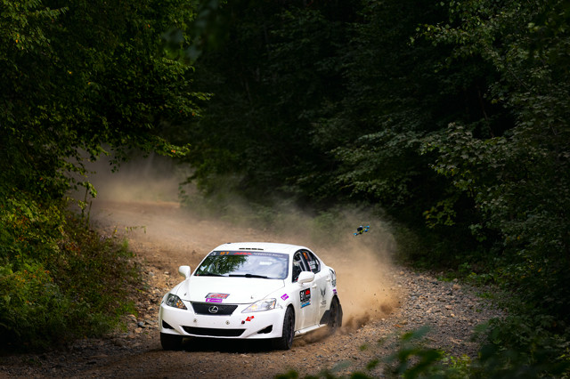 A white Lexus IS250 snakes through the forest at speed, being chased by a drone.