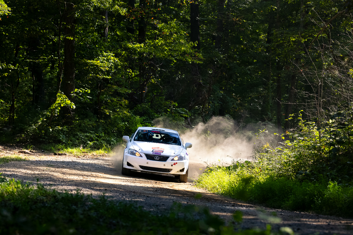 A white Lexus IS250 races around a corner in the woods with a dust trail.