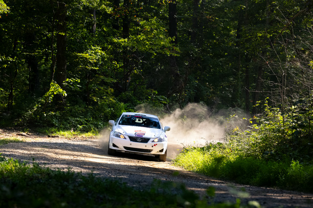 A white Lexus IS250 races around a corner in the woods with a dust trail.