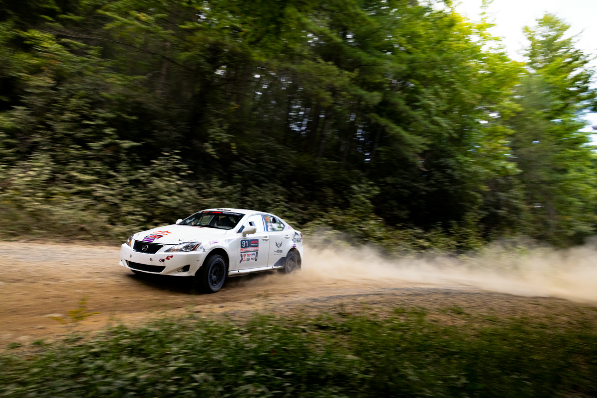 A white Lexus IS250 powering through a dirt turn in the woods with a dust trail behind it.