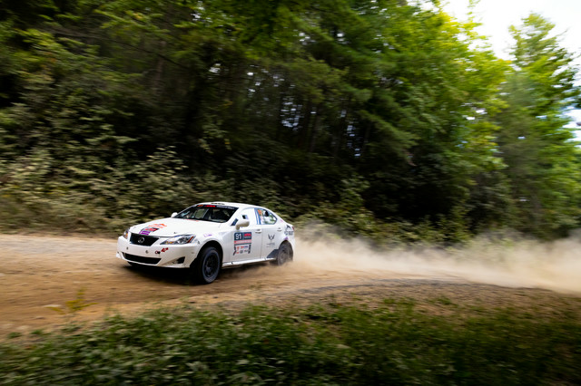 A white Lexus IS250 powering through a dirt turn in the woods with a dust trail behind it.