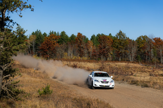 A white Lexus IS250 hauling down a dirt road, carrying some angle while a massive dust cloud can be seen behind it.